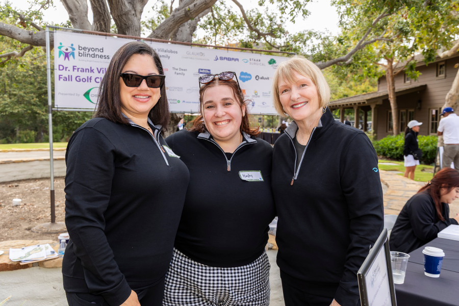 Three woman smiling in front of a banner and trees.
