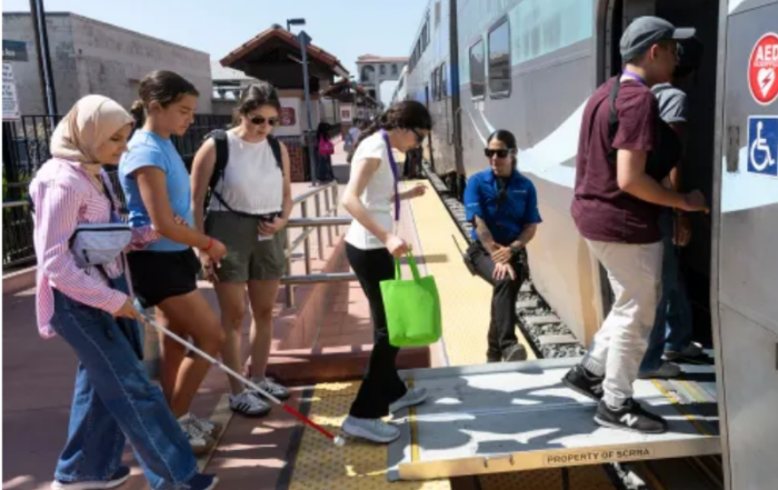 Seven people walking onto a train. One holding a white cane.